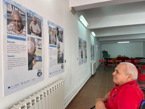 Utente homem da Unidade de Cuidados Continuados Integrados de Bragança, no museu etnográfico da Santa Casa, a contemplar o seu cartaz com fotografias e textos dele transcritos de uma entrevista que lhe foi feita com frases respeitantes à sua educação e ao seu trabalho.